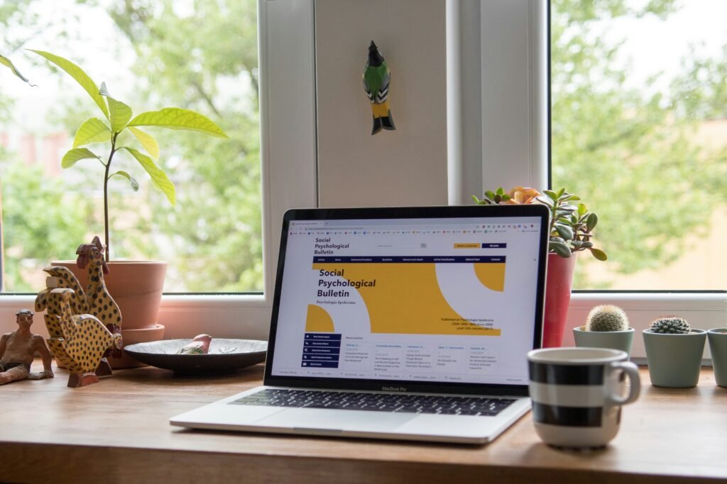 A laptop on a wooden desk displays a website. The desk also holds potted plants, a ceramic rooster, a small giraffe figurine, a plate, and a striped mug. There’s a window with greenery outside in the background.